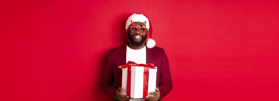 Christmas. Handsome african american man in party glasses and santa hat holding new year gift, bring present in box and smiling, standing over red background.