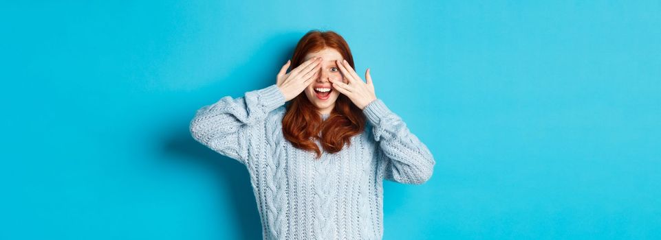 Excited teenage redhead girl open eyes to see holiday surprise, receiving presents, looking amazed at camera, standing over blue background.