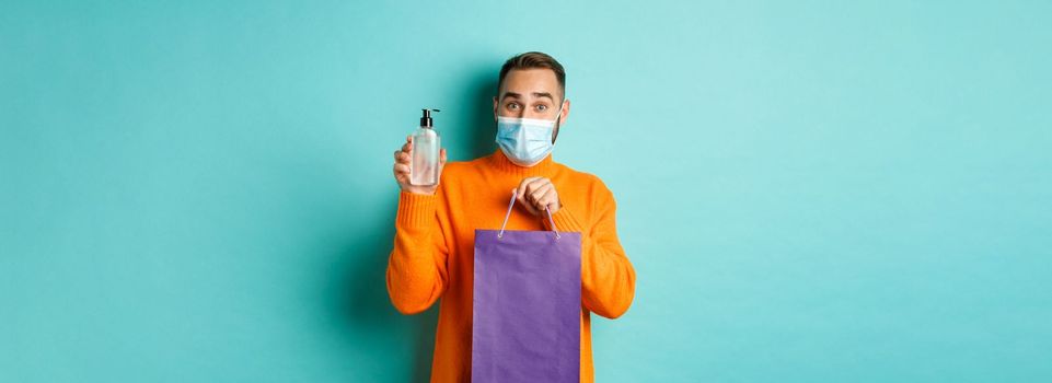 Coronavirus, pandemic and lifestyle concept. Man in face mask showing shopping bag and hand sanitizer, standing over turquoise background.