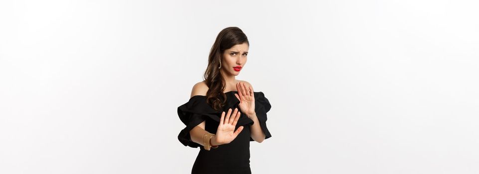 Fashion and beauty. Reluctant and worried woman asking to stay away, showing stop gesture and looking scared, standing in black dress over white background.