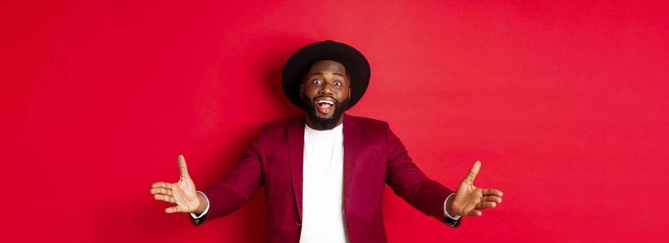 Fashion and party concept. Pleased handsome Black man spread hands in welcome gesture, reaching to receive something, looking amazed, standing over red background.