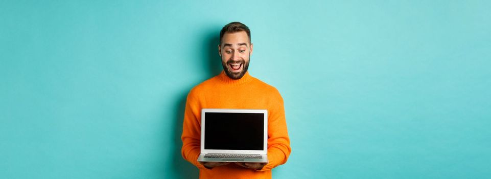 Excited adult male model showing laptop screen, demonstrating online promo, standing happy against light blue background.