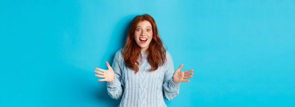 Excited redhead woman shaking hands, explaining big news, staring at camera amazed, standing against blue background.