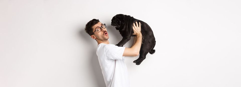 Image of handsome young man loving his pug. Dog owner holding puppy and smiling happy at camera, standing over white background.
