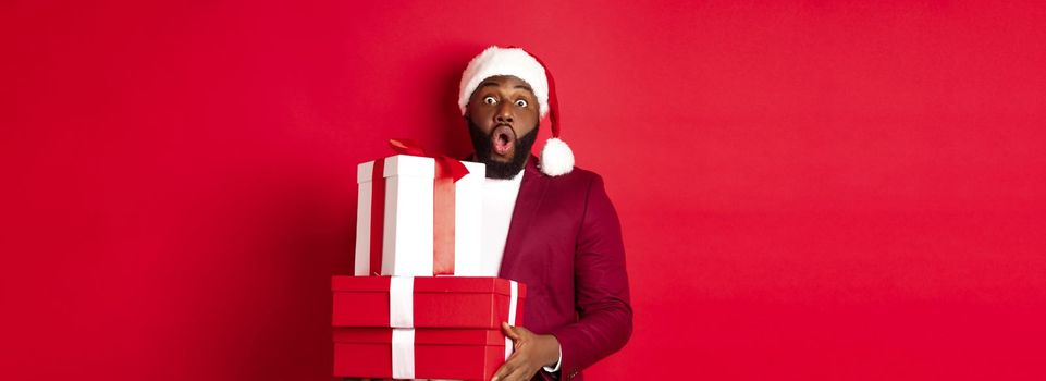 Christmas, New Year and shopping concept. Cheerful Black man secret santa holding xmas presents and smiling excited, bring gifts, standing against red background.