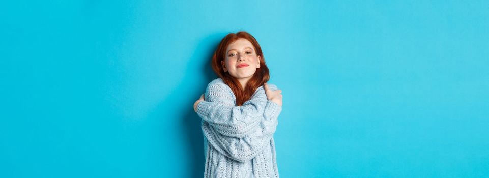 Happy cute redhead girl hugging herself, wearing comfortable and warm sweater, smiling at camera, standing over blue background.