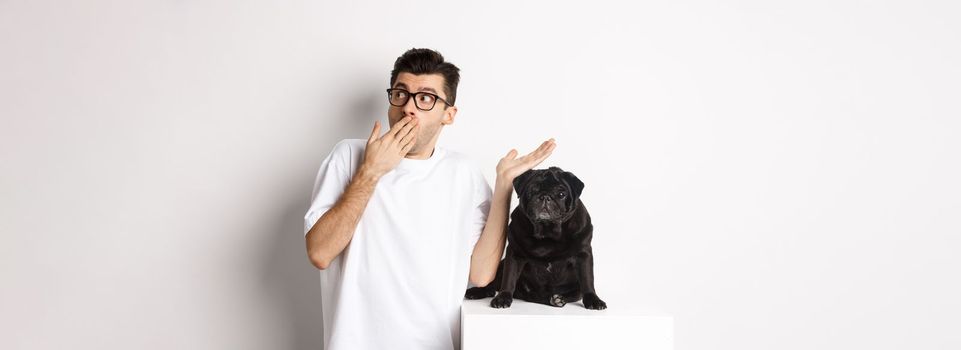 Surprised and shocked man in glasses, standing near cute black dog and staring right at logo, posing over white background.