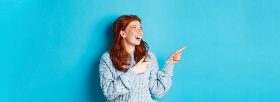 Excited redhead girl in sweater, looking and pointing fingers left, showing promo offer or logo, standing over blue background.