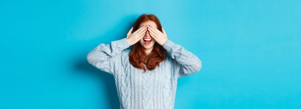 Cheerful redhead female model close eyes and waiting for christmas gift, holding hands on face and smiling amused, anticipating surprise, standing over blue background.