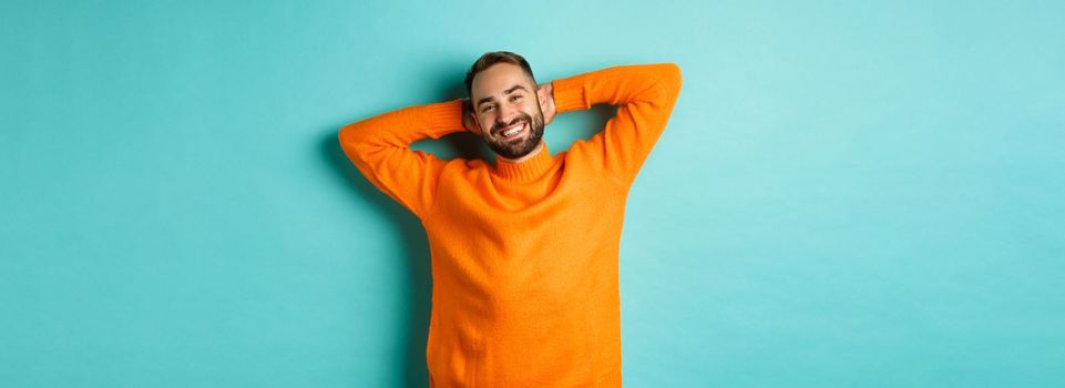 Handsome man relaxing, resting and smiling, holding hands behind head with carefree face, standing against light blue background.