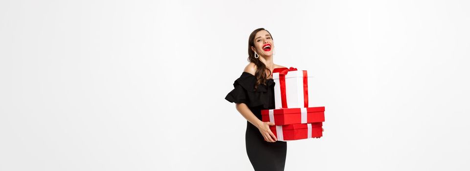 Merry christmas and new year holidays concept. Full length of attractive woman in elegant dress laughing, holding xmas gifts, laughing happy, white background.