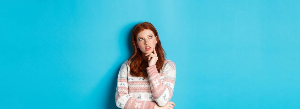 Image of thoughtful redhead girl looking at upper left corner, thinking or making choice, standing in winter sweater over blue background.