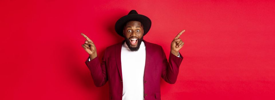 Winter holidays and shopping concept. Cheerful Black man smiling and showing two promo, pointing fingers sideways at copy spaces, red background.