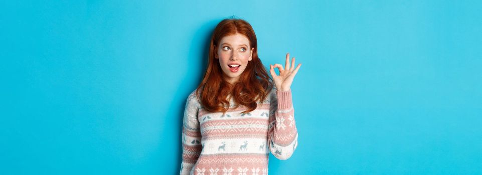 Silly redhead woman in sweater, showing okay sign and staring at upper left corner logo, standing over blue background satisfied.