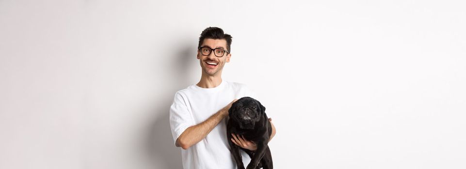 Happy hipster guy in glasses pet dog and smiling. Cute black pug enjoy spending time with owner, looking satisfied, standing over white background.