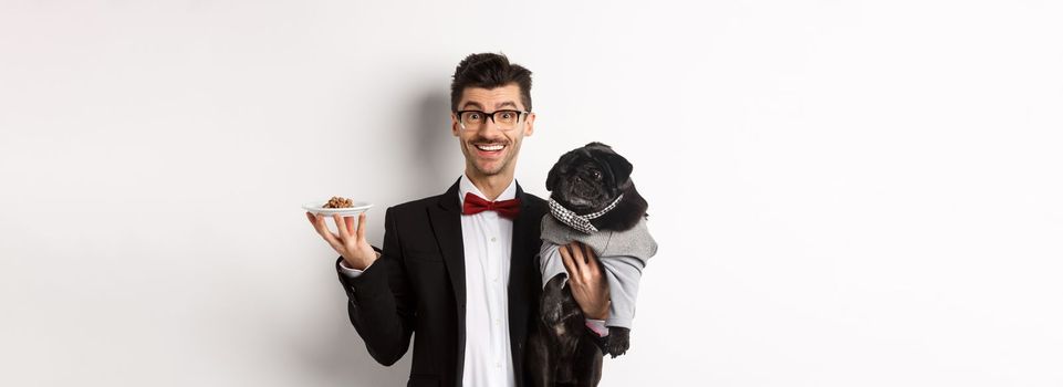 Handsome young hipster in suit and glasses holding cute black pug and pet food on plate, standing over white background.