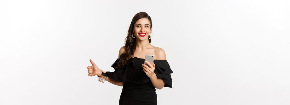 Online shopping concept. Attractive woman in trendy black dress, makeup, showing thumb-up and using mobile phone app, white background.