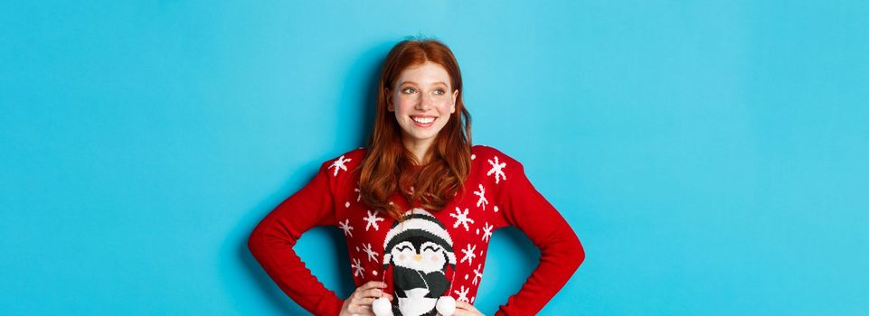Winter holidays and Christmas Eve concept. Beautiful teenage redhead girl in xmas sweater looking left at logo, smiling pleased, holding hands on waist, blue background.