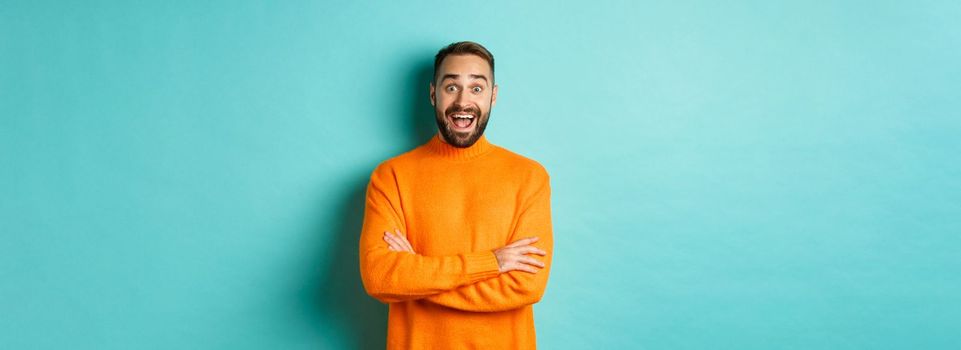 Image of happy and surprised man reacting to news, looking amazed, standing in orange sweater against turquoise background.