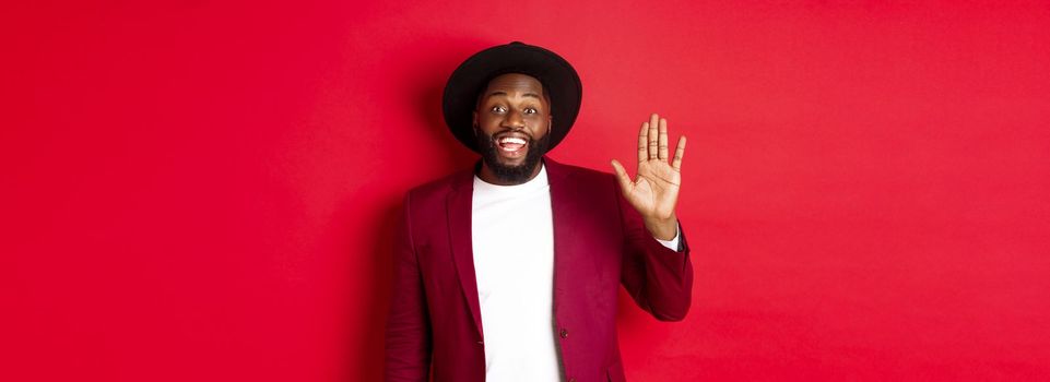 Friendly african american guy waving hand, saying hello and smiling, greeting you, standing over red background.