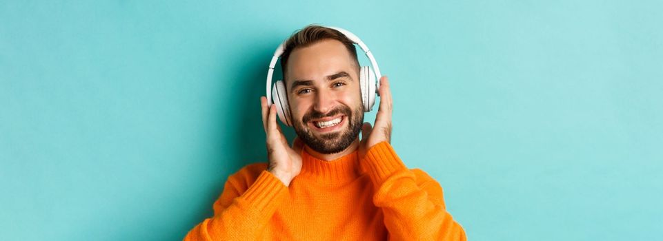 Close-up of handsome modern man listening music in headphones, standing in orange sweater over turquoise background.