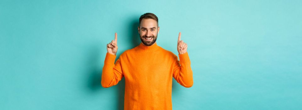 Handsome bearded guy in orange sweater showing up promo, pointing at top and smiling, standing over turquoise background.