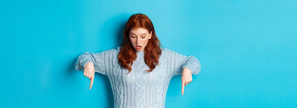 Winter holidays and people concept. Impressed redhead girl in sweater, looking and pointing down with amazement, standing against blue background.