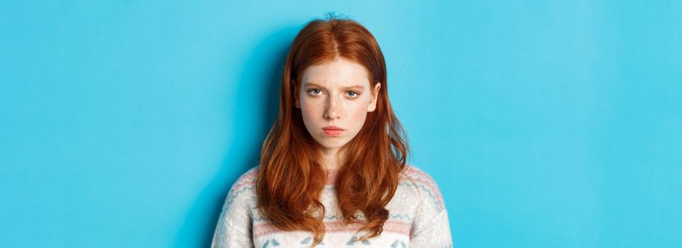 Close-up of angry redhead teenage girl looking pissed-off at camera, frowning and sulking mad, standing over blue background.