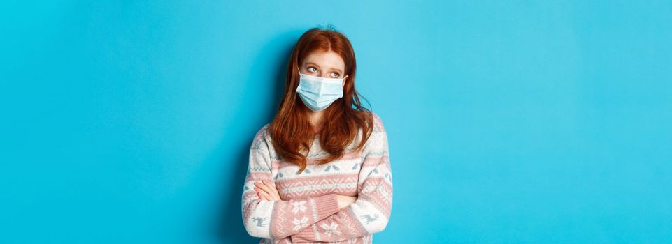 Winter, covid-19 and pandemic concept. Ignorant redhead teenage girl in face mask, roll eyes and looking unamused, cross arms on chest reluctant, standing over blue background.