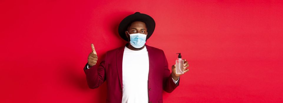 Covid-19, quarantine and holidays concept. Young african american man in medical mask, showing hand sanitizer and thumb up, recommending to use antiseptic.