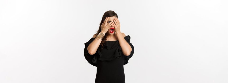 Fashion and beauty. Shocked young woman in black dress covering eyes, peeking through fingers at something embarrassing, cringe, standing over white background.