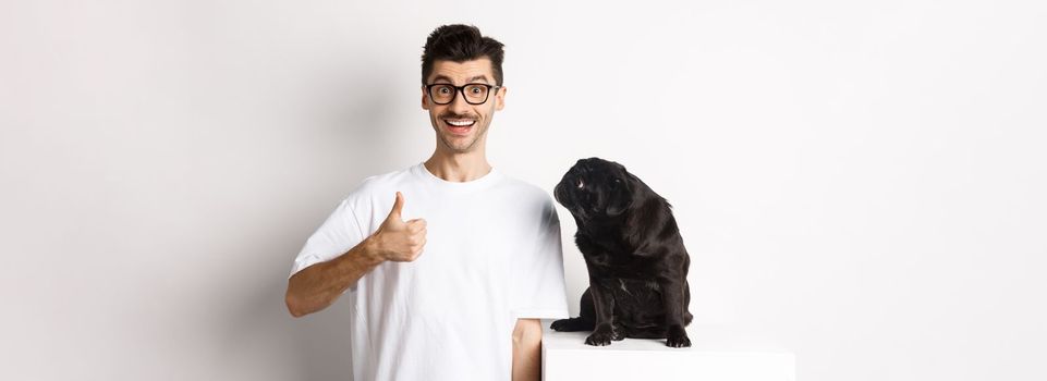 Cheerful handsome guy standing near cute black pug and showing thumb-up. Pet owner approve and recommend product for dogs, white background.