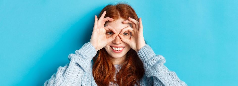 Close-up of funny and cute redhead girl making hand glasses and looking through them, seeing promo offer and smiling, standing over blue background.