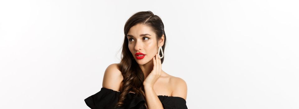 Fashion and beauty concept. Close-up of elegant woman in black dress, showing earrings and looking sensual, red lipstick and makeup on, white background.