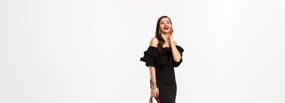 Beauty and fashion concept. Full length of beautiful young lady going on party in heels, black dress and makeup, holding elegant purse, laughing at camera, white background.