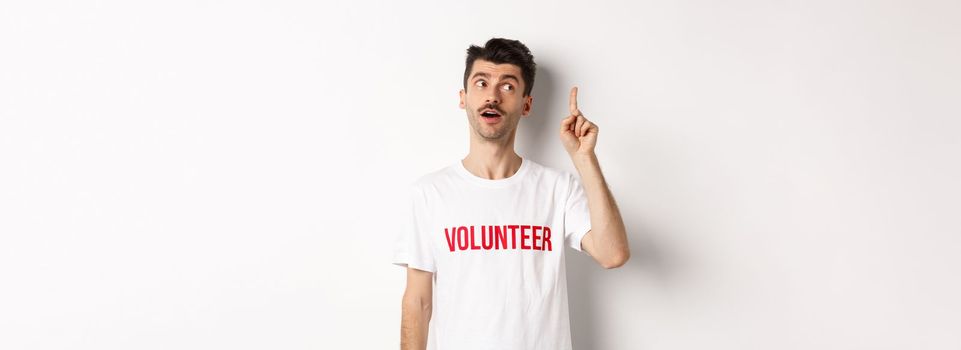 Handsome young man in volunteer t-shirt having an idea, raising finger and saying suggestion, white background.