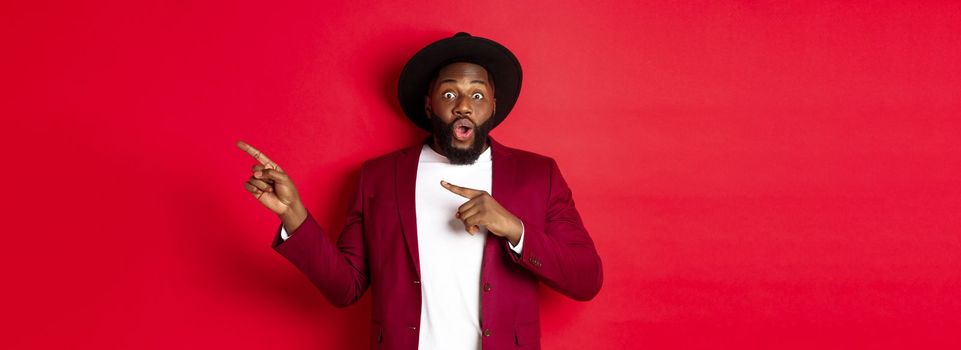 Winter holidays and shopping concept. Stylish african american man pointing fingers right at copy space for logo, standing over red background.