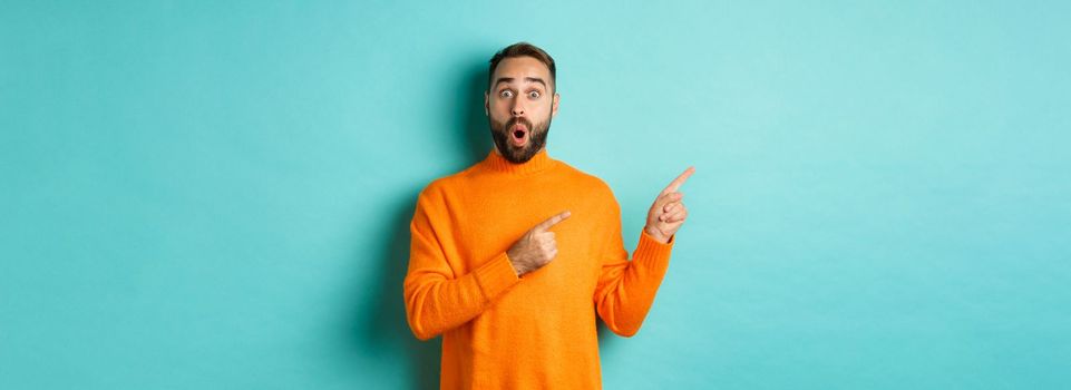 Impressed young man with beard, reacting to promo banner, pointing fingers left, showing logo and looking surprised, standing over turquoise background.