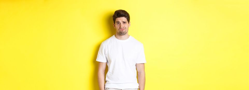 Disappointed guy looking upset, sulking and frowning, standing displeased against yellow background.