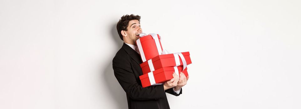 Concept of christmas holidays, celebration and lifestyle. Image of excited handsome man buying gifts for new year, standing happy over white background in black suit.