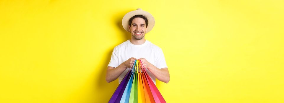 Image of happy man shopping on vacation, holding paper bags and smiling, standing against yellow background.