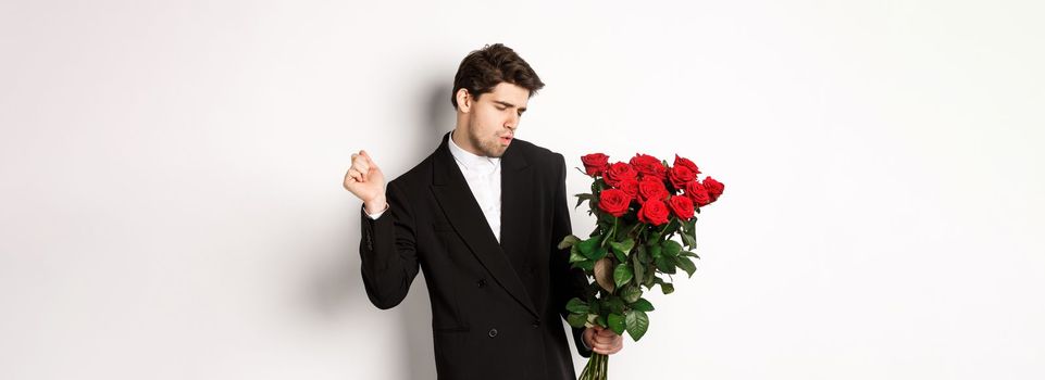 Image of elegant and sassy man in black suit, looking confident and holding bouquet of red roses, going on a romantic date, standing against white background.