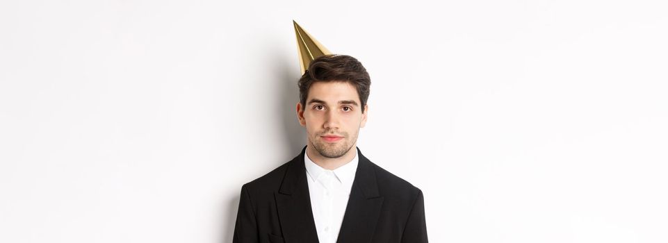 Close-up of handsome man in party hat and trendy suit, celebrating new year, standing against white background.