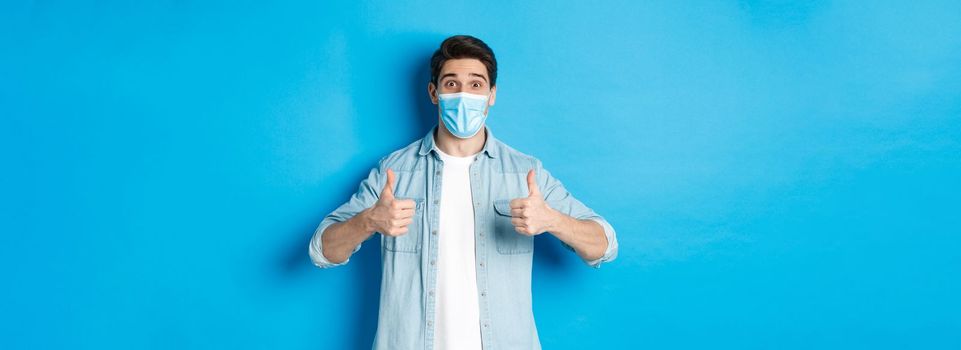 Concept of covid-19, pandemic and social distancing. Amazed guy in medical mask showing thumbs-up, recommending promo offer, standing against blue background.