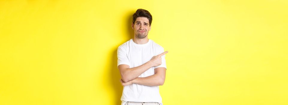 Skeptical and reluctant guy in white t-shirt, pointing finger left and smirking unamused, showing bad advertisement, standing over yellow background.