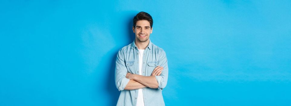 Handsome guy in casual clothes standing with arms crossed and confident smile against blue background.