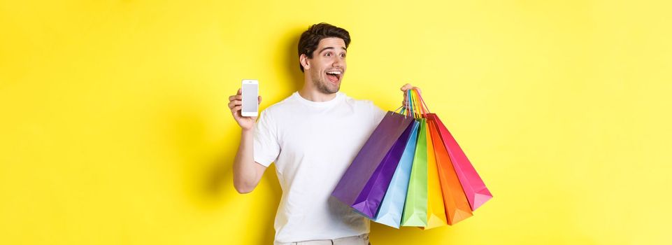 Excited man showing smartphone screen and shopping bags, achieve app goal, demonstrating mobile banking application, yellow background.