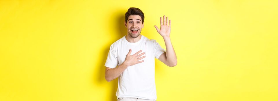 Happy guy making promise, holding hand on heart, swearing to tell truth, standing over yellow background in white t-shirt.