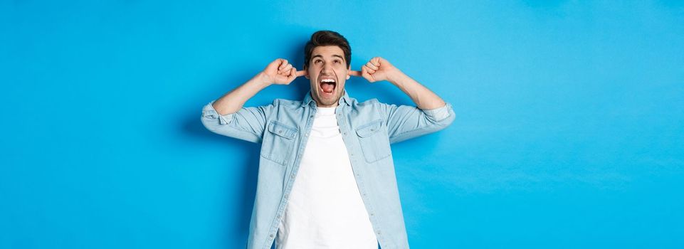 Irritated man close ears from noisy neighbours, looking up and shouting annoyed, hearing loud music, standing over blue background.