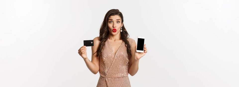 Online shopping and holidays concept. Excited beautiful lady in elegant dress showing mobile screen and credit card, standing over white background.